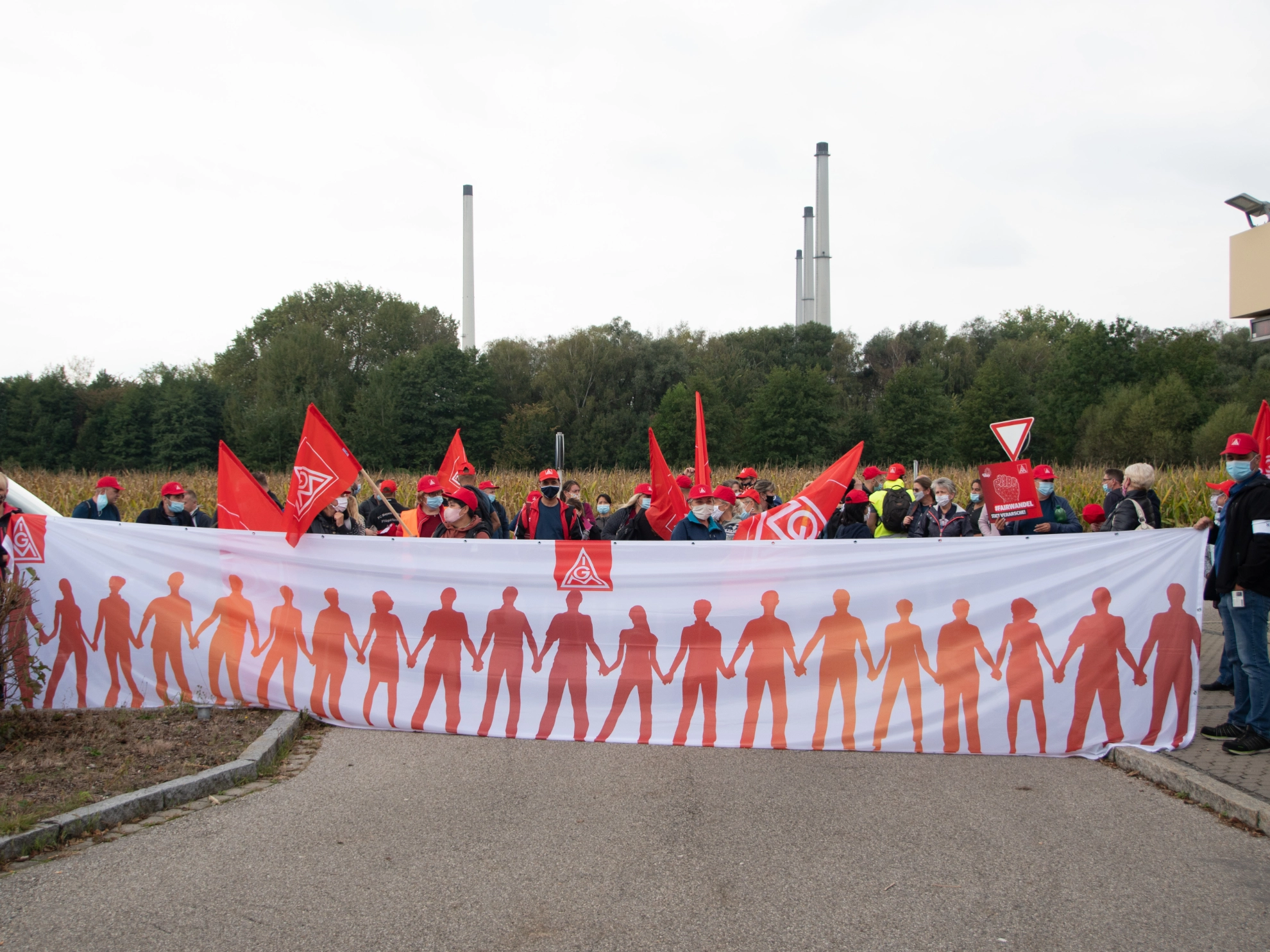 Kolleginnen und Kollegen mit IG Metall Fahnen und Mützen, mit Transparent, wo sich Menschen an der Hand halten. 