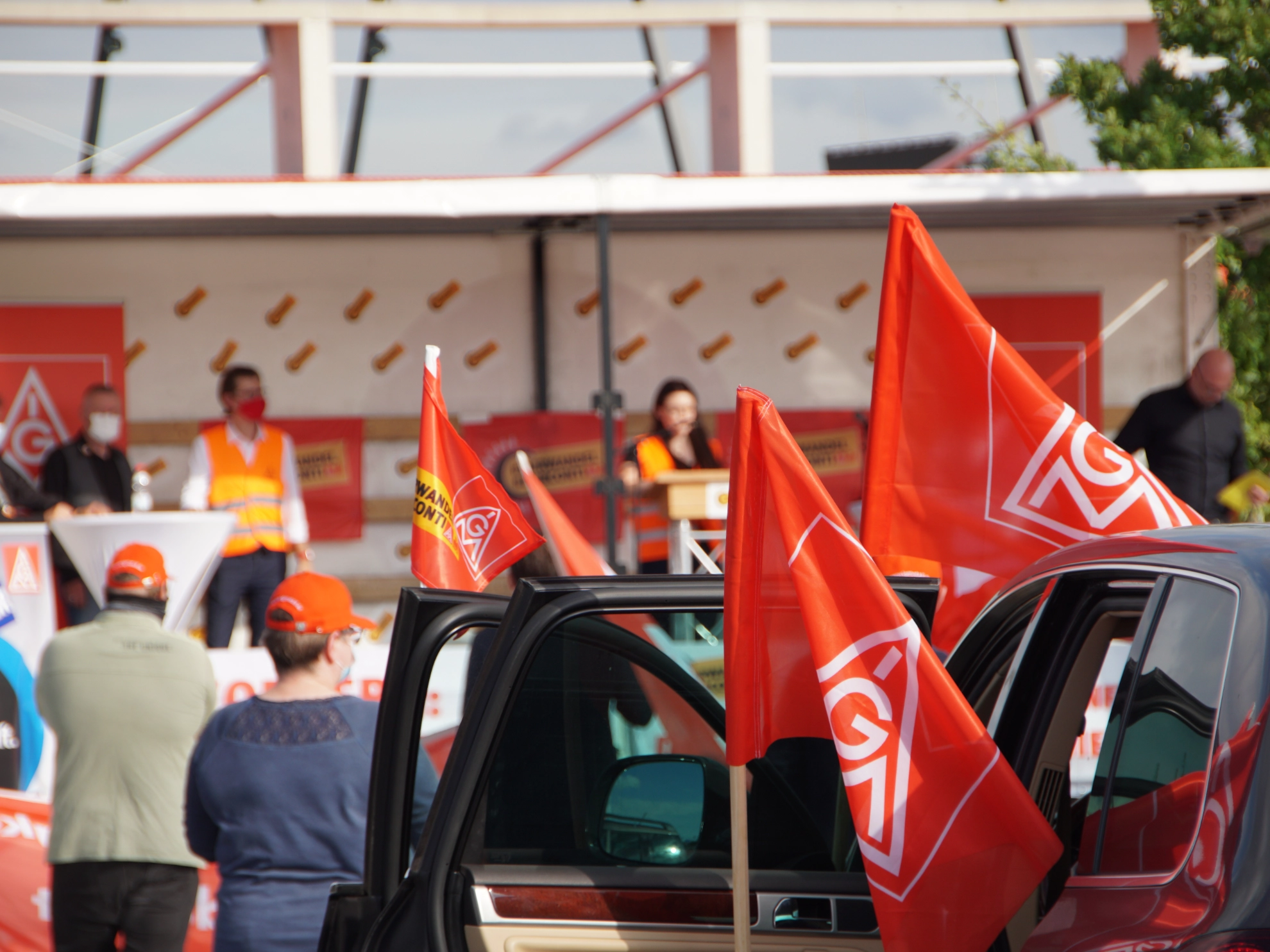 Blick auf die Bühne, Rednerin und Kollegen auf der Bühne unscharf, Autos und Fahnen im Vordergrund