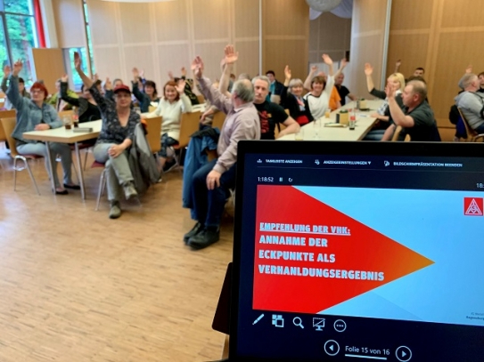 Kolleginnen und Kollegen die die Hand heben für eine Abstimmung in einem Saal. Im Vordergrund ein Monitor mit IGM Vorlage zur Abstimmung. 